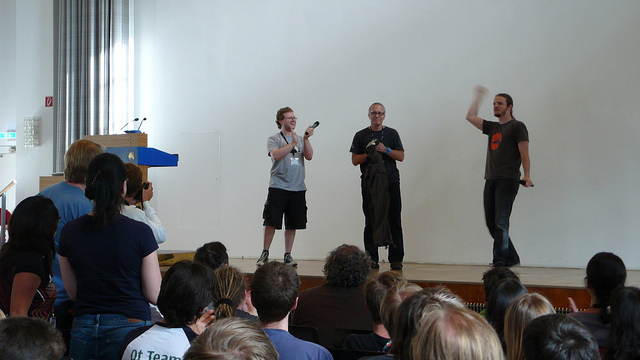 Matthias standing on a stage, he's in-between two people. He's facing a big audience in a closed room